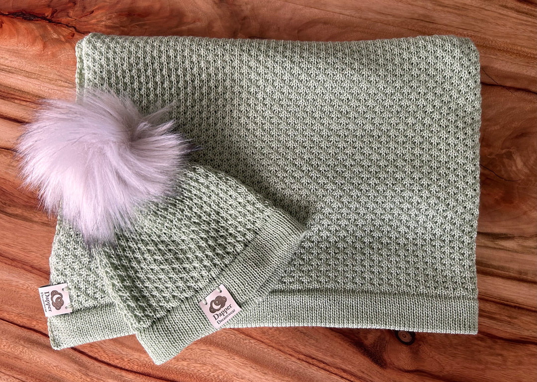 A sage green coloured textured merino wool baby blanket and beanie with a pom pom. Photographed on a wooden table background. Made in Australia from Australian grown wool. 